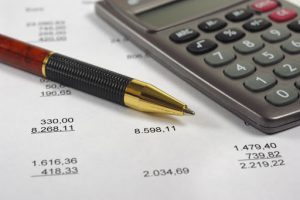 Close-up of a calculator and a pen resting atop a paper budget with various lines of expenses and income.